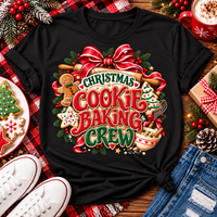 A black short-sleeve graphic T-shirt laid flat on a rustic wooden surface with holiday props. The shirt features a large festive cookie-baking crew design with red ribbon, green pine branches, gingerbread cookies, a Christmas tree, and the text 'Christmas Cookie Baking Crew.