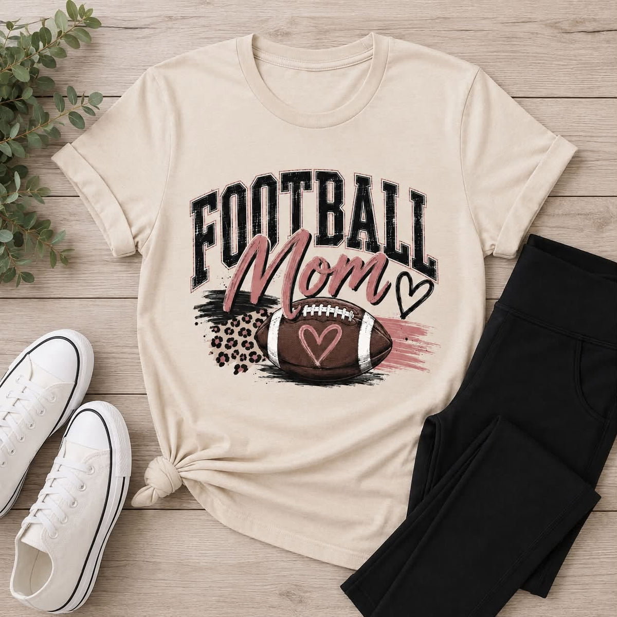 Cream Football Mom graphic T-shirt with distressed black lettering, blush pink accents, leopard print details, and a football with hearts.