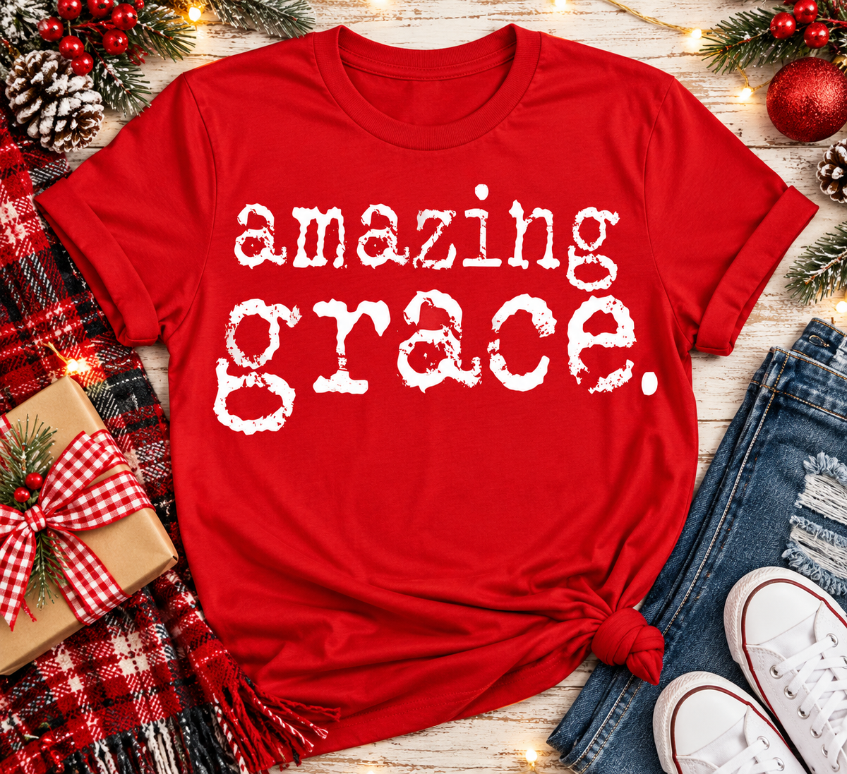 A red short-sleeve graphic T-shirt with the phrase 'amazing grace.' in distressed white lettering, styled in a holiday flat lay with jeans, white sneakers, a plaid blanket, pinecones, and Christmas decorations.