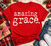 A red short-sleeve graphic T-shirt with the phrase 'amazing grace.' in distressed white lettering, styled in a holiday flat lay with jeans, white sneakers, a plaid blanket, pinecones, and Christmas decorations.