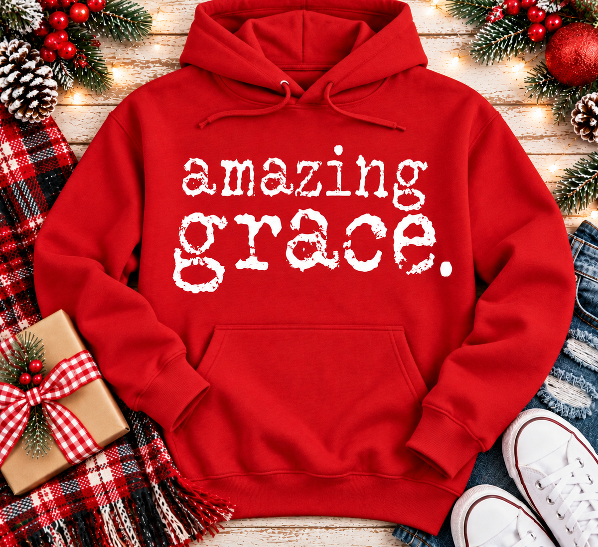 A red pullover hoodie with the words 'amazing grace.' printed in white distressed lettering across the chest, styled in a holiday flat-lay with a plaid scarf, gift box, pinecones, ornaments, jeans, and white sneakers on a wooden surface.