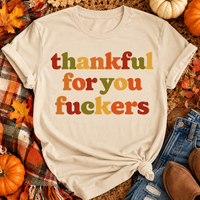 Funny Thankful for You Fuckers Thanksgiving graphic tee featuring bold retro lettering in burnt orange, mustard yellow, olive green, and rust red. Women's fall-inspired short sleeve crewneck shirt available in beige and brown, perfect for Thanksgiving and Friendsgiving.
