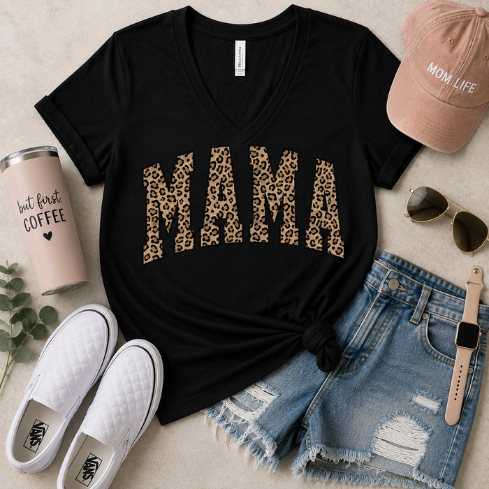 A styled flat-lay featuring a black V-neck t-shirt with 'MAMA' printed in leopard-print letters, surrounded by white slip-on sneakers, distressed denim shorts, a pink 'MOM LIFE' cap, sunglasses, a watch, and a coffee cup on a light beige background.