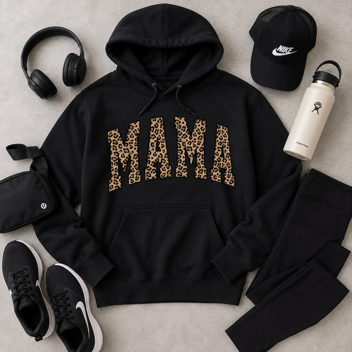 A black pullover hoodie with the word 'MAMA' printed across the chest in leopard-print letters, styled in a flat lay with a black cap, white water bottle, black headphones, black crossbody bag, black sneakers, and black leggings on a light gray surface.