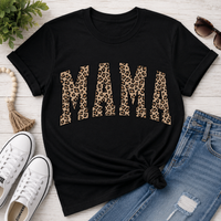 Black graphic t-shirt featuring bold leopard print "MAMA" lettering, styled with distressed blue jeans, white sneakers, sunglasses, and neutral accessories for a casual everyday mom outfit.