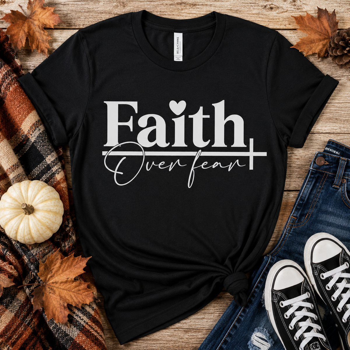 Black Faith Over Fear graphic T-shirt styled in a fall flat lay with jeans, sneakers, a plaid blanket, autumn leaves, and a white pumpkin.