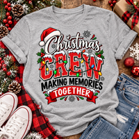 Christmas Crew Making Memories Together graphic on a pink crewneck T-shirt, featuring red lettering, colorful Christmas lights, a Santa hat, holly, and snowflakes.