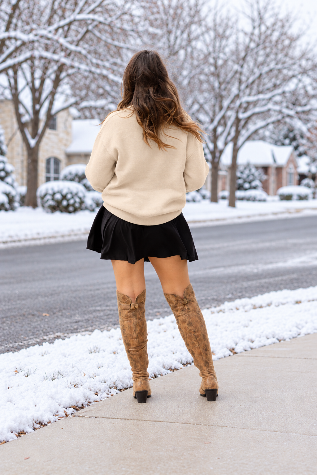 A woman stands on a snowy sidewalk wearing a cream crewneck sweatshirt with a holiday tree graphic and the words 'Merry and Bright', a black pleated mini skirt, and tan knee-high boots.