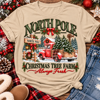 North Pole Christmas Tree Farm vintage holiday graphic shirt featuring a red pickup truck carrying Christmas trees, snowy evergreen trees, rustic farmhouse scenery, festive red bows, and distressed green lettering reading North Pole Christmas Tree Farm Always Fresh. Available in beige and black in short sleeve crewneck, V-neck, long sleeve crewneck, and hoodie styles.