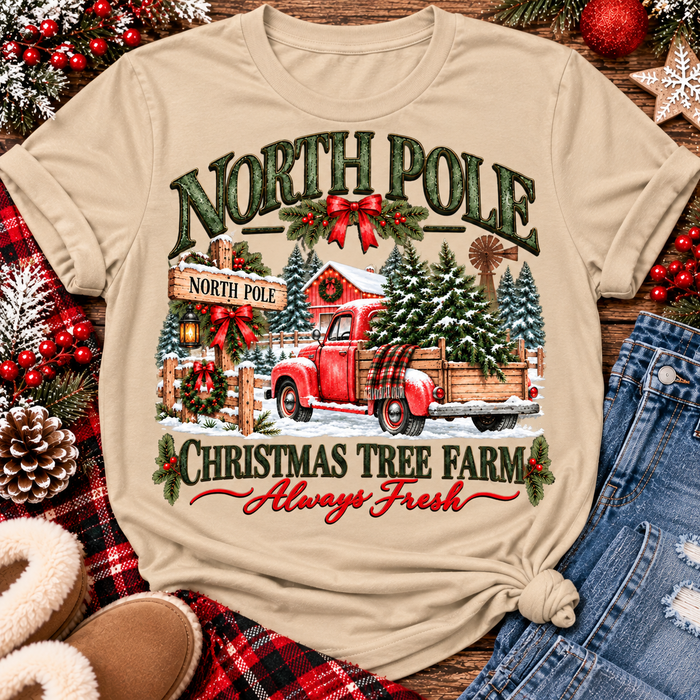 North Pole Christmas Tree Farm vintage holiday graphic shirt featuring a red pickup truck carrying Christmas trees, snowy evergreen trees, rustic farmhouse scenery, festive red bows, and distressed green lettering reading North Pole Christmas Tree Farm Always Fresh. Available in beige and black in short sleeve crewneck, V-neck, long sleeve crewneck, and hoodie styles.