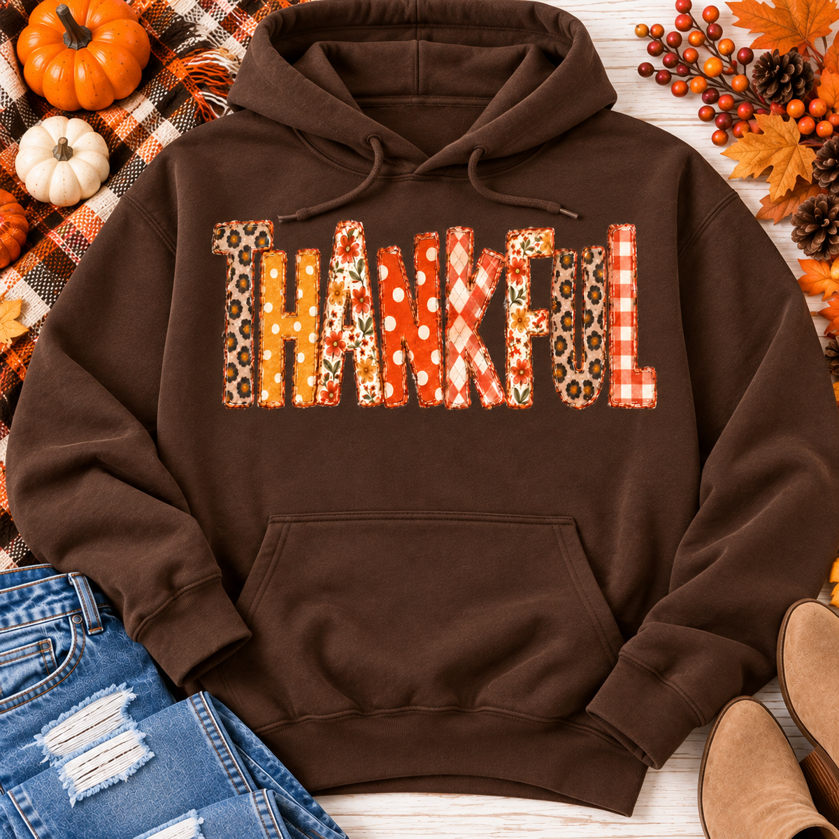 Brown Thankful fall graphic hoodie with orange floral, plaid and polka dot patchwork lettering, styled with jeans and autumn decorations.