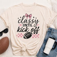 Cream crewneck Classy Until Kickoff football graphic tee featuring a pink bow, black football, pink checkerboard pattern and black and pink lettering.