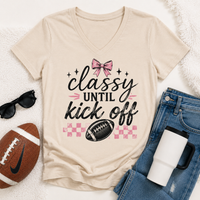 Cream V-neck Classy Until Kickoff football graphic tee featuring a pink bow, black football, pink checkerboard pattern and black and pink lettering.