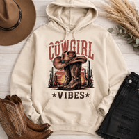 Cream Cowgirl Vibes hoodie featuring vintage cowboy boots, western cowgirl hat, cactus and desert sunset graphic in rustic brown and red tones.