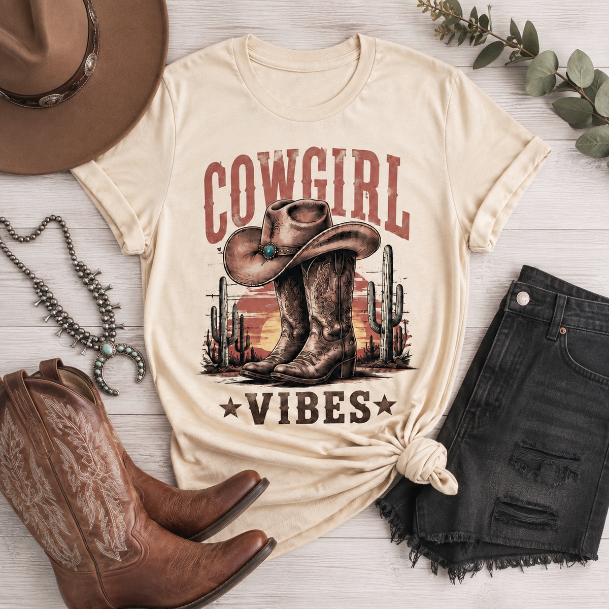 Cream Cowgirl Vibes graphic tee featuring vintage cowboy boots, western hat, cactus and desert sunset design in rustic brown and red tones.