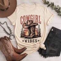 Cream Cowgirl Vibes graphic tee featuring vintage cowboy boots, western hat, cactus and desert sunset design in rustic brown and red tones.
