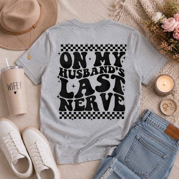 Gray crewneck graphic tee featuring oversized black “On My Husband’s Last Nerve” lettering with retro checkerboard details on the back.